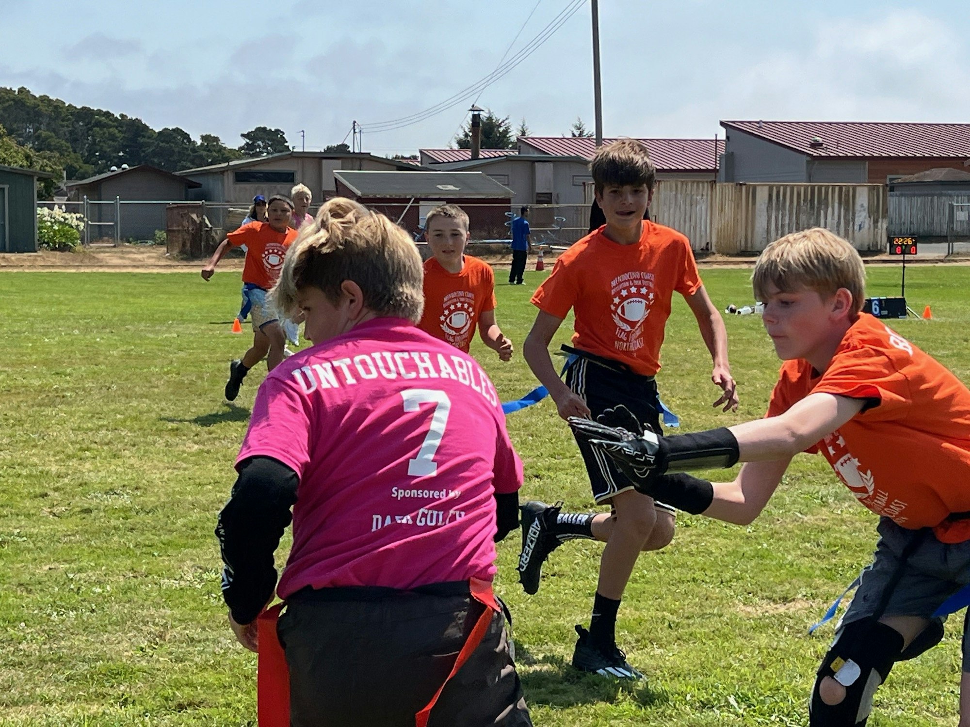 Children playing tag football outdoors with one wearing a pink jersey and others in orange.