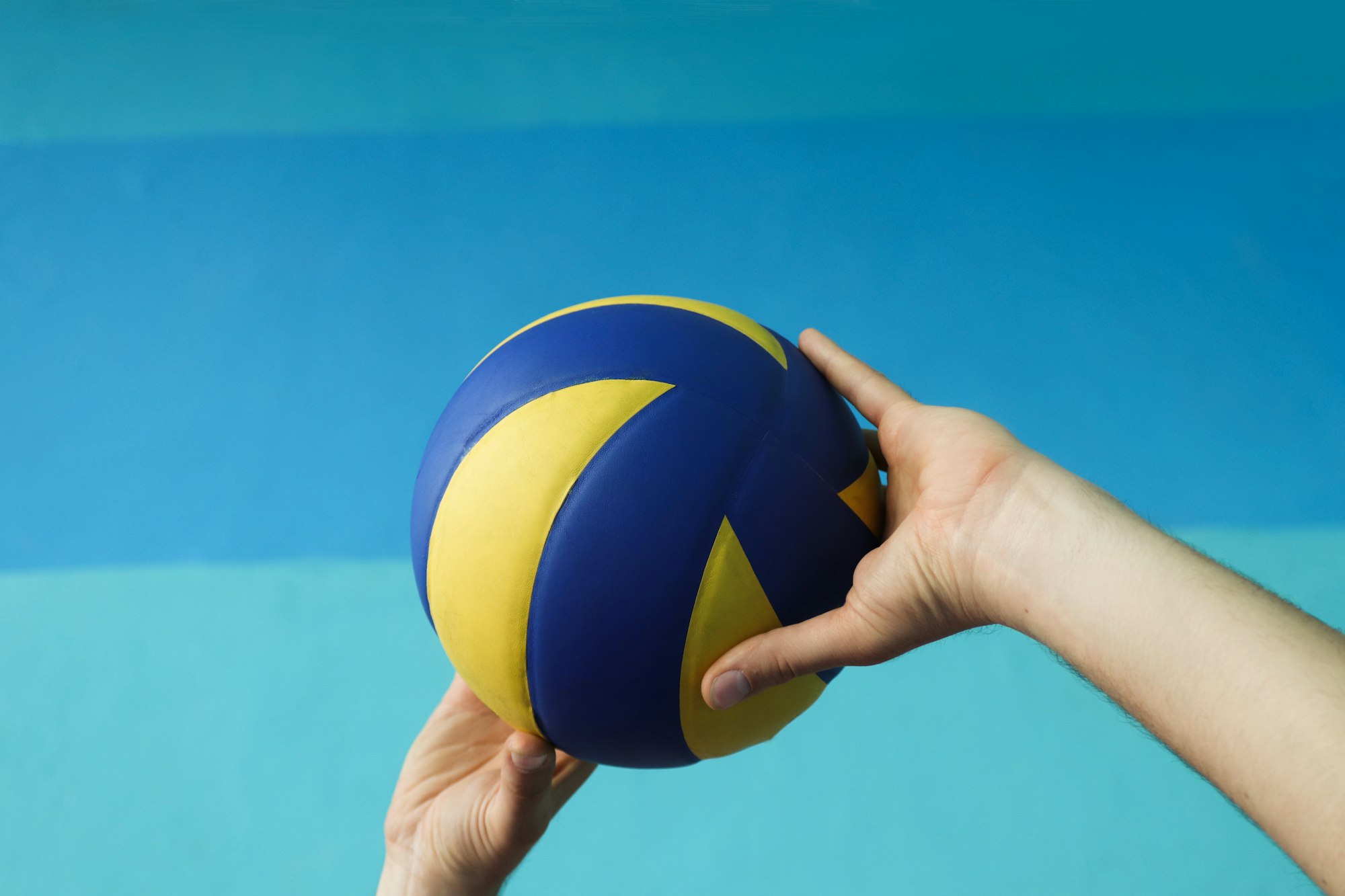 A hand holding a blue and yellow volleyball against a blue background.