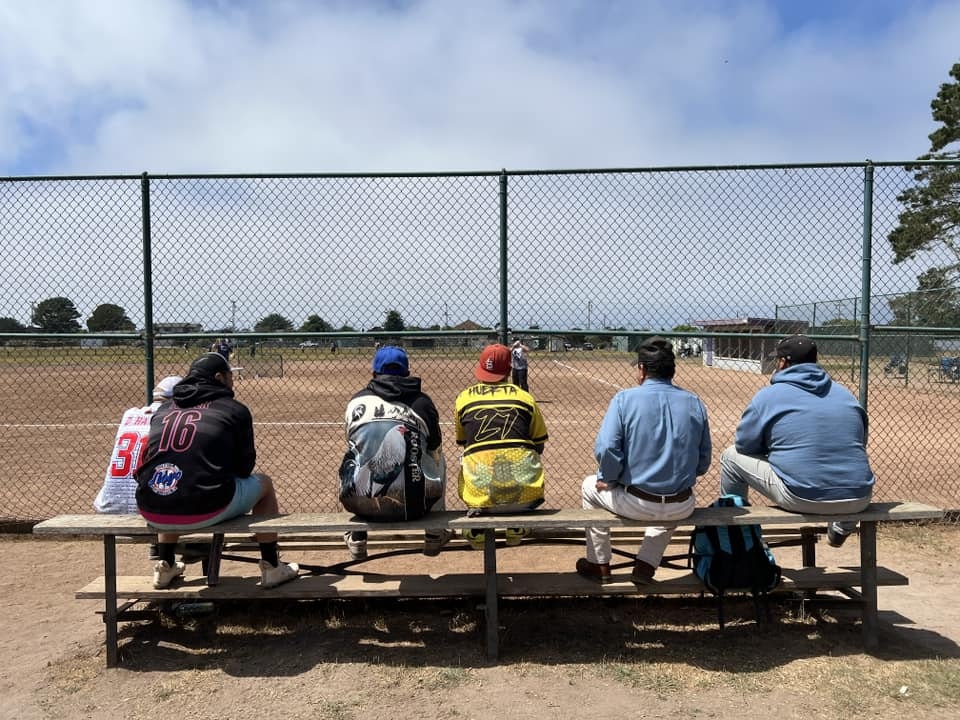 People sitting on a bench watching a game, separated by a chain-link fence from a sports field.