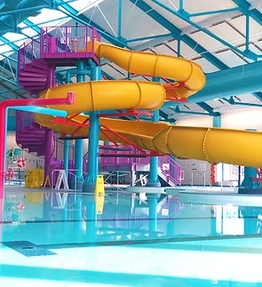 Indoor water park scene with a large yellow and orange spiral slide above a clear blue pool.
