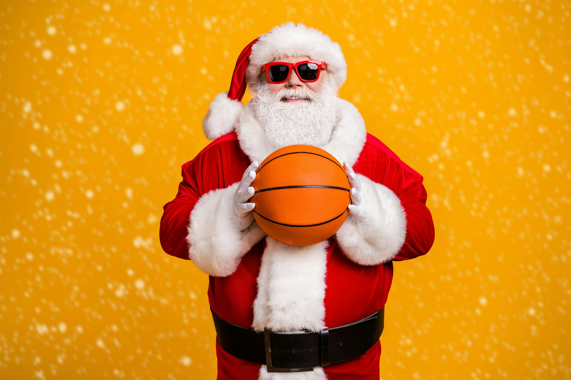Santa wearing sunglasses holding a basketball against a yellow snowy background.