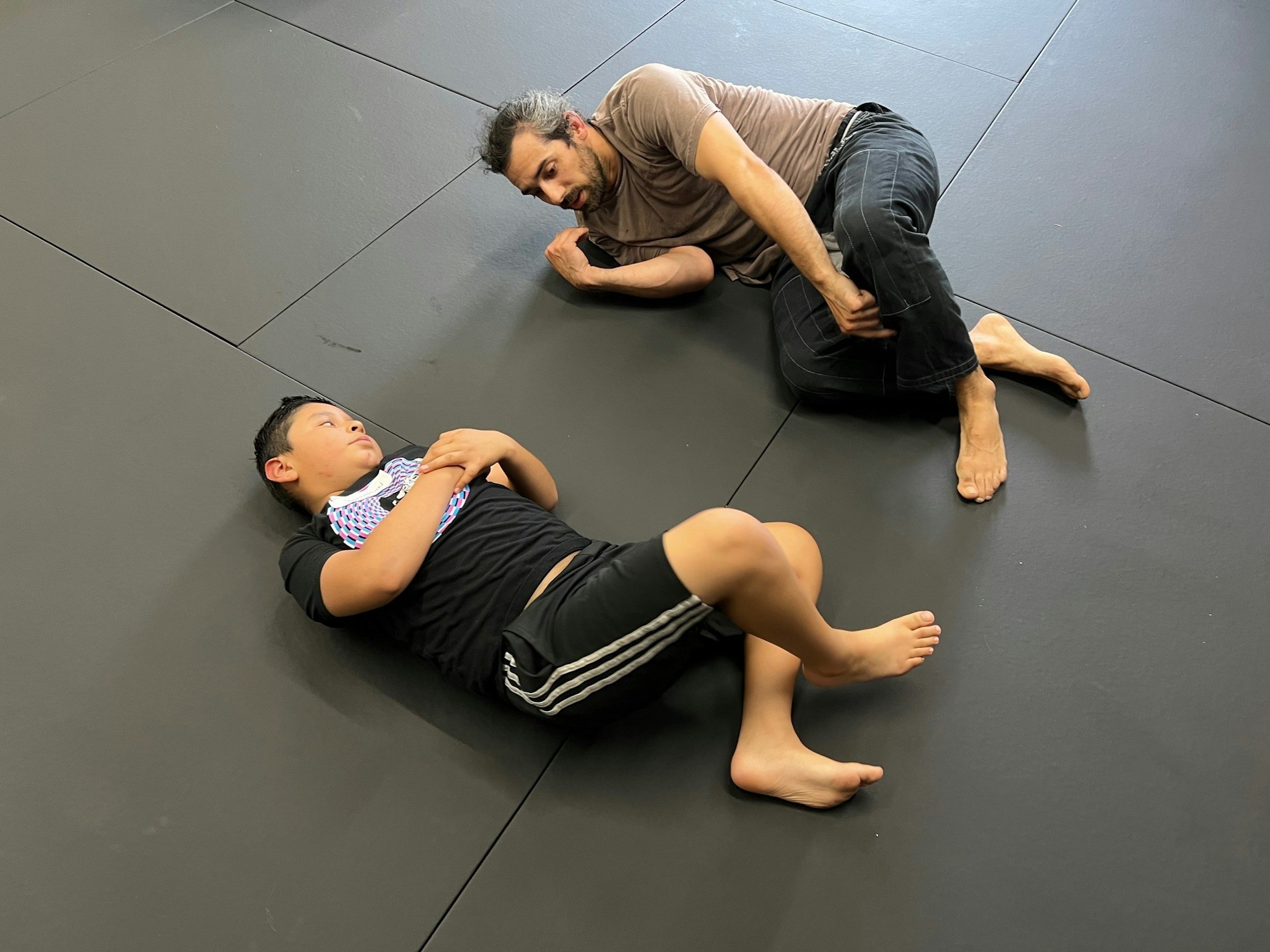 Two individuals on a mat, one standing, one lying down, possibly in a martial arts training session.