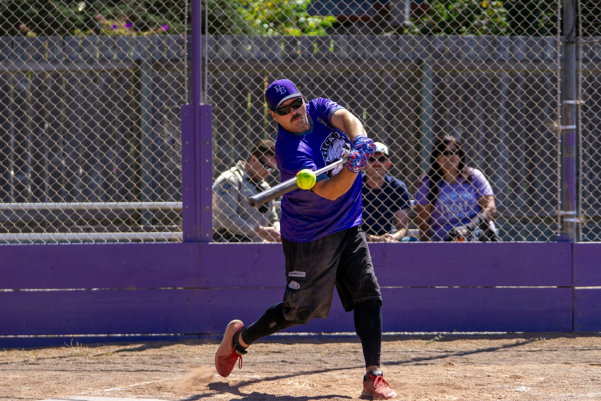 Person hitting a softball with a bat on a field, wearing a purple shirt and cap, with spectators behind a fence.