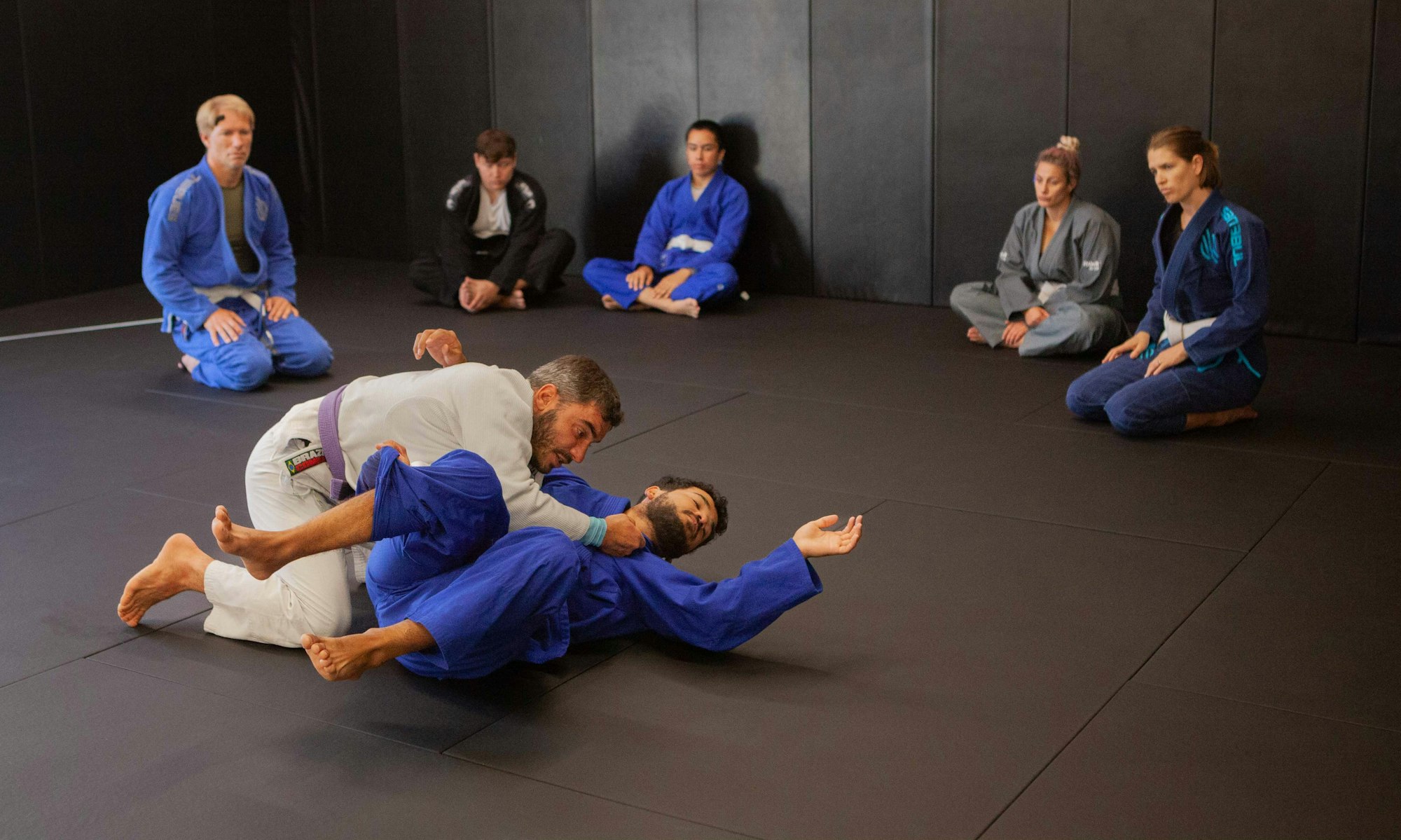 Two people practicing Jiu-Jitsu on a mat while others watch.