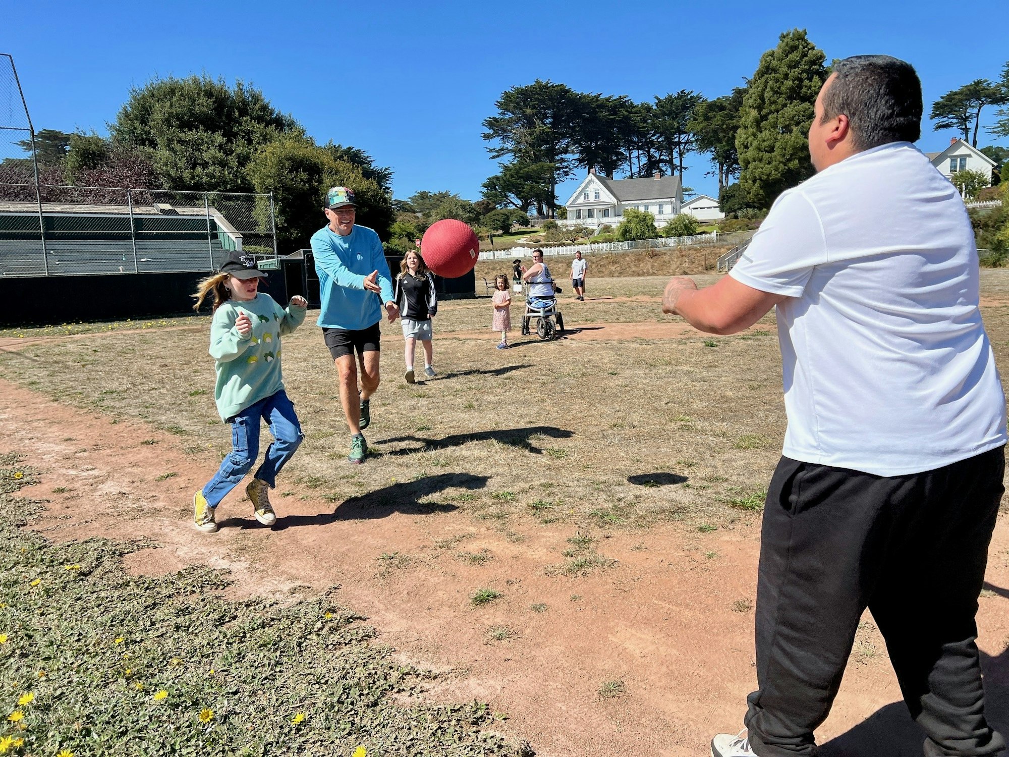 People playing with a large red ball outdoors on a sunny day, with trees and houses in the background.
