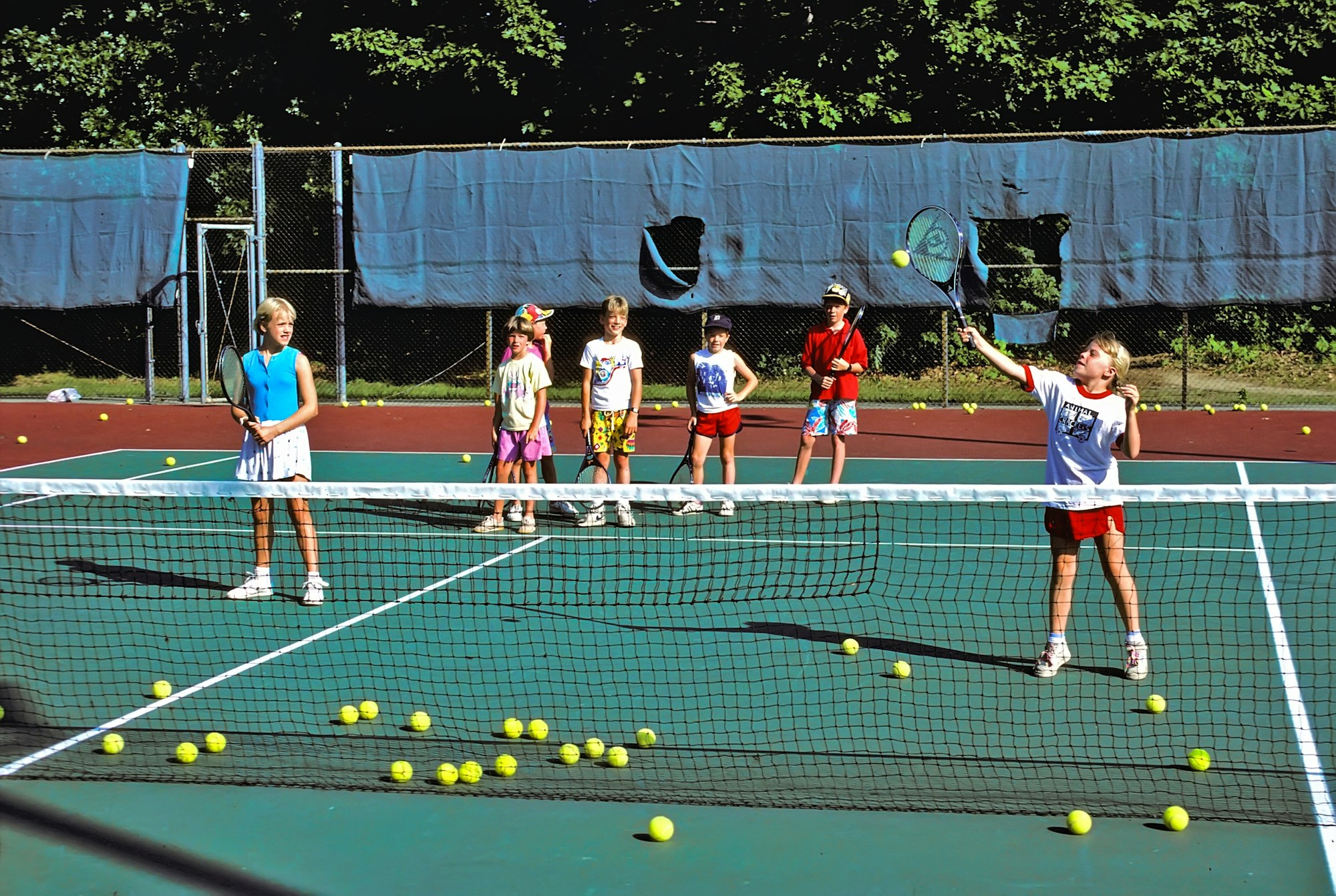 Kids on a tennis court practicing, with scattered yellow tennis balls and one child hitting a ball.