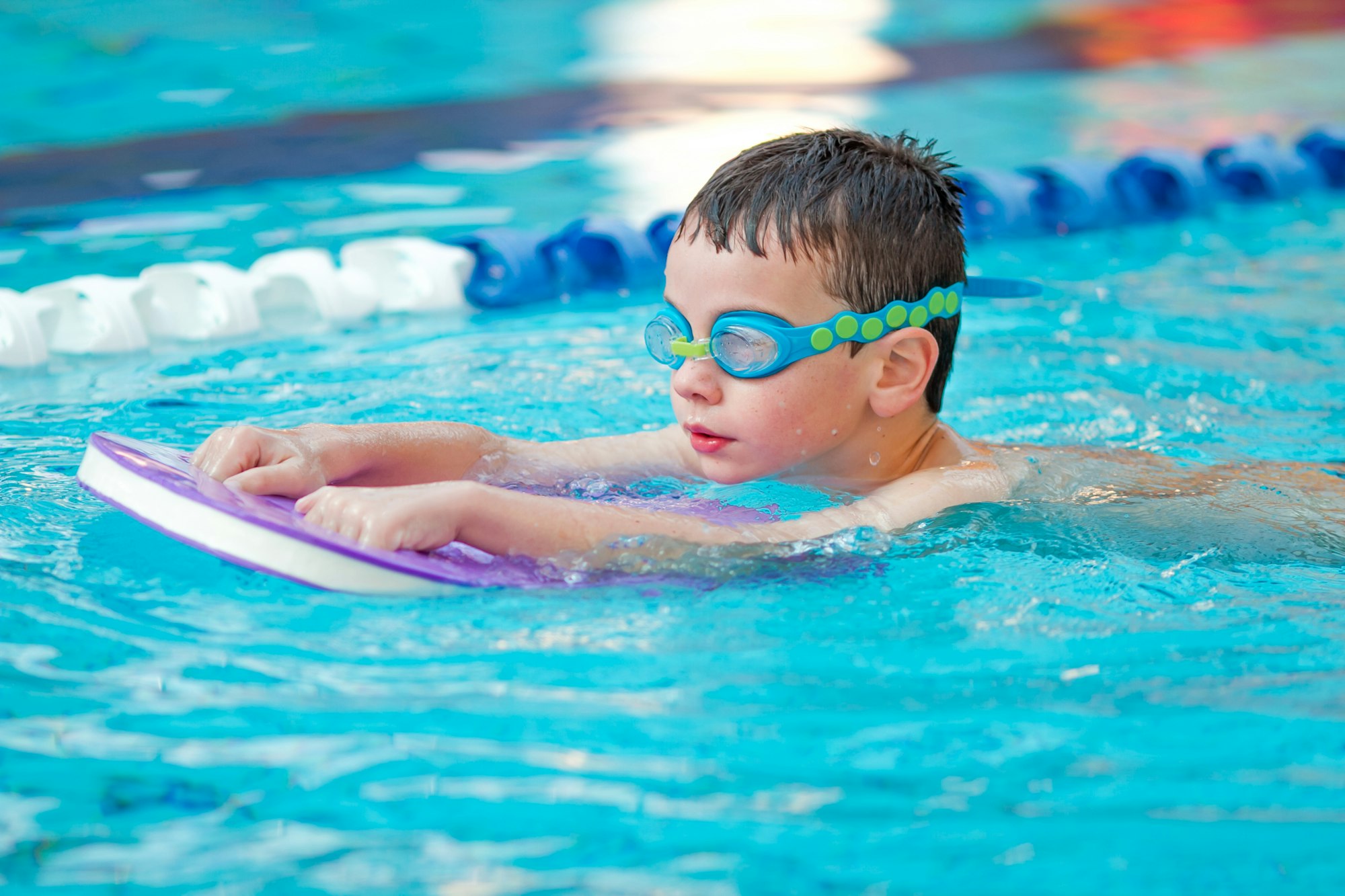 A child with goggles floats on a kickboard in a swimming pool.