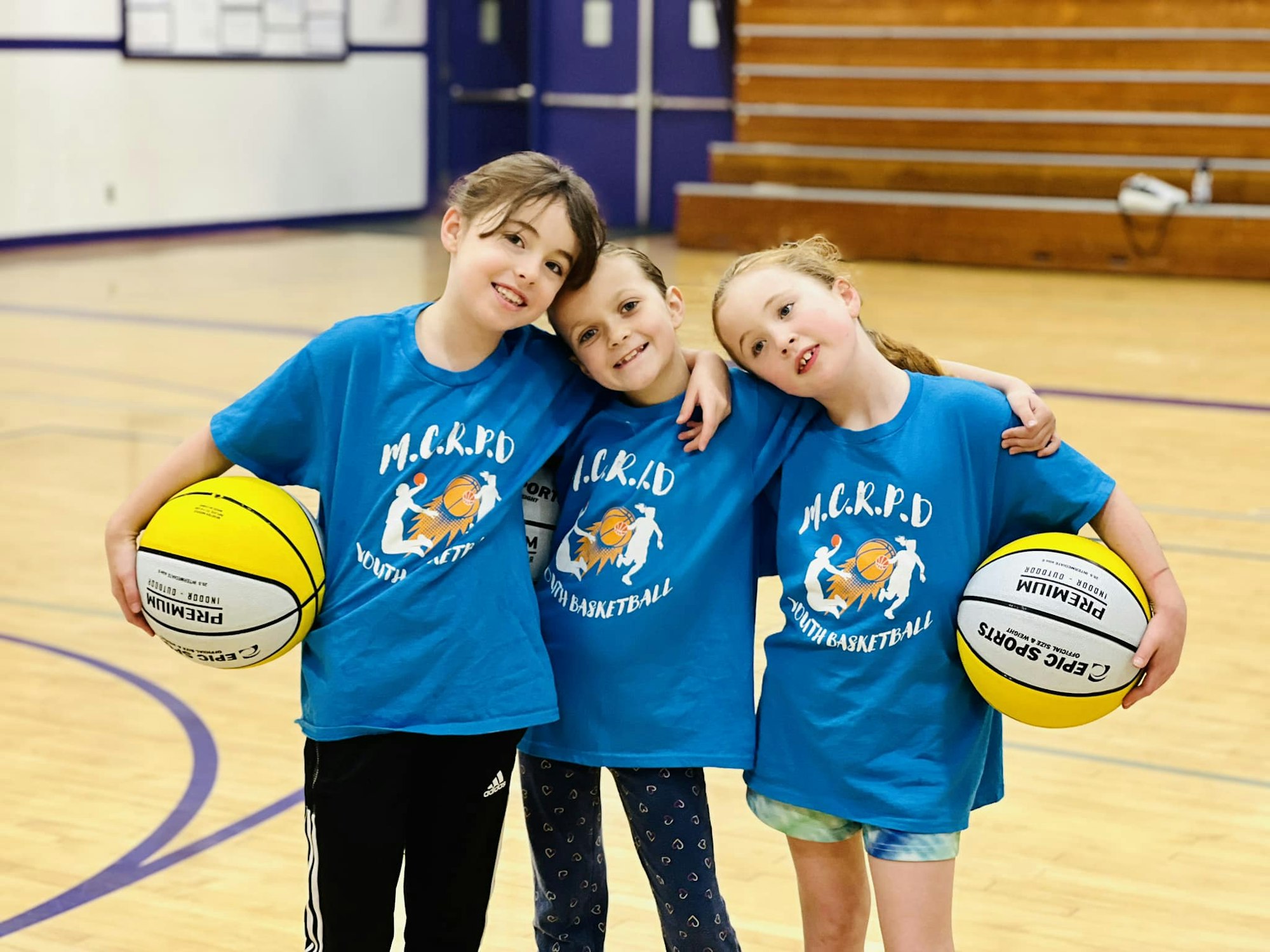 Three children in blue sports shirts, holding basketballs, smiling in a gymnasium.