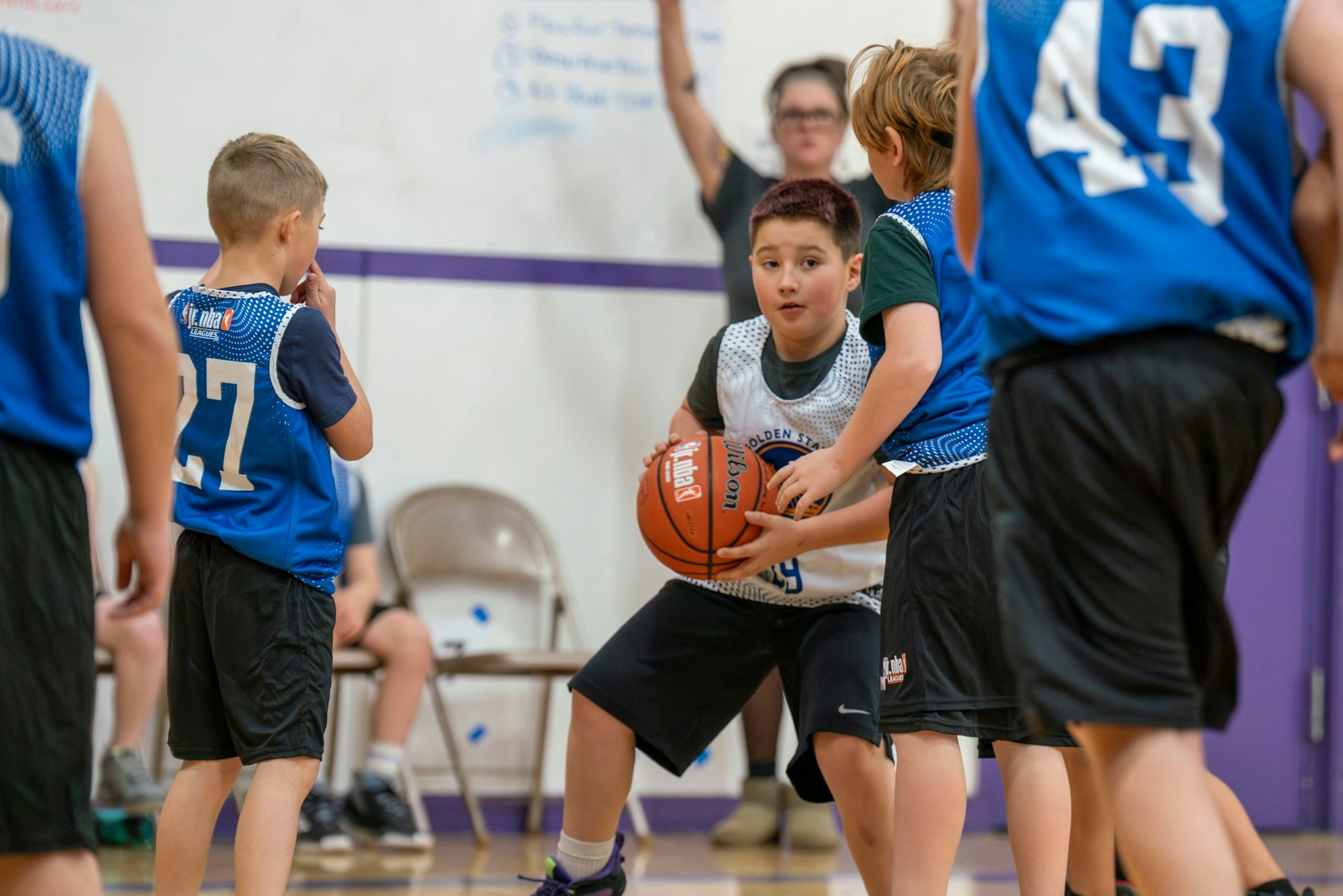 Kids playing basketball indoors, one boy holding the ball while others in blue jerseys defend him.