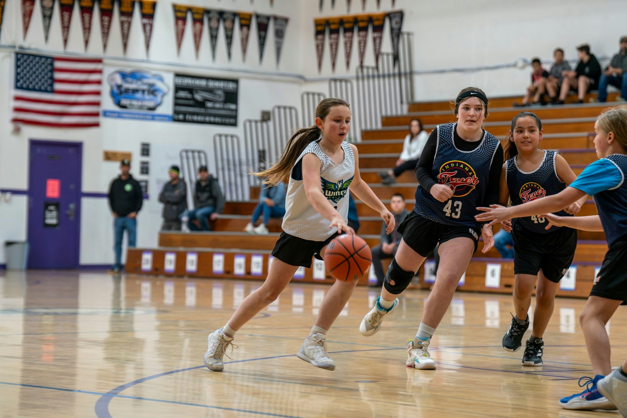 Girls playing basketball in a gym; one dribbling past defenders. Spectators and flag in background.