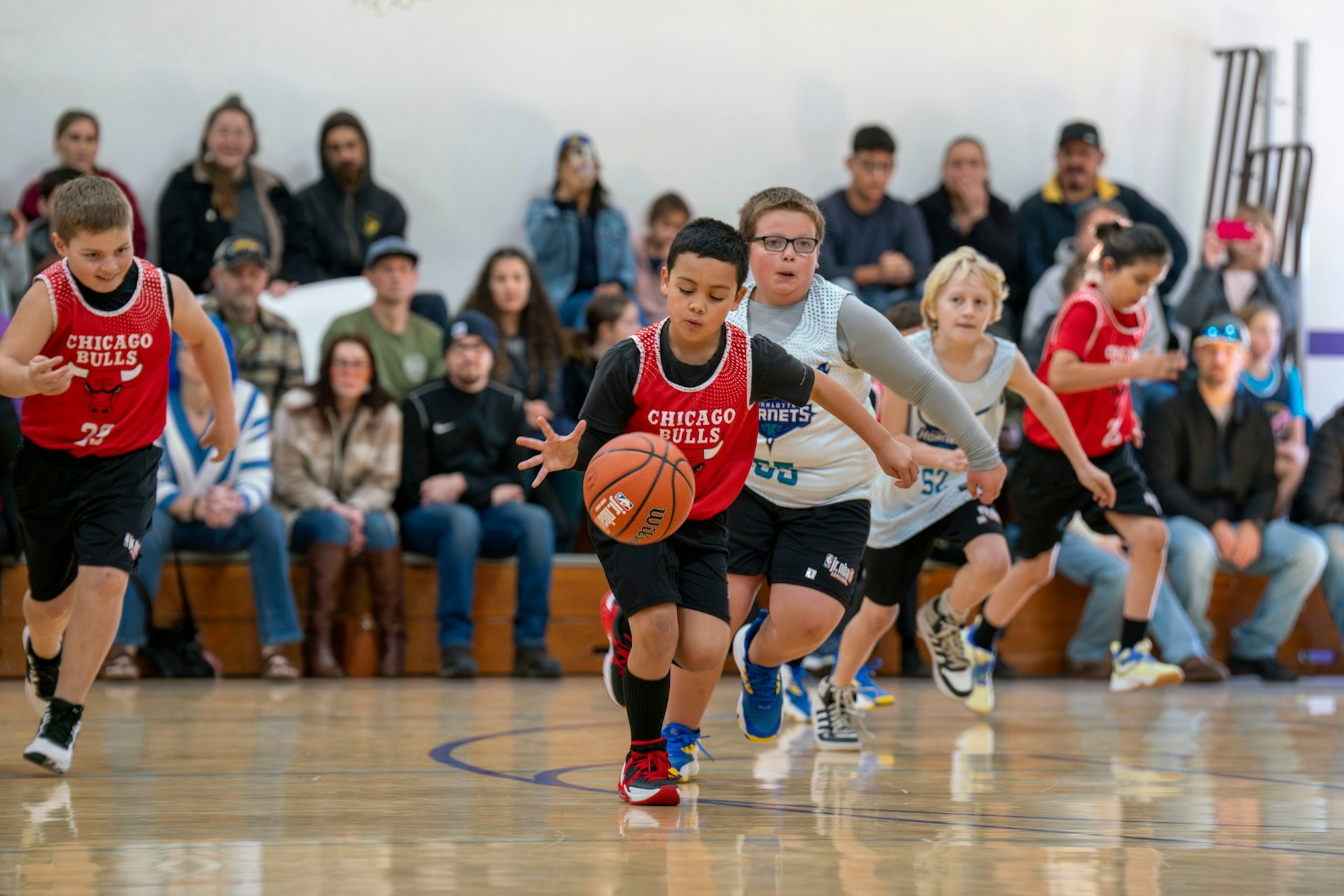 Kids playing basketball, one in a red jersey dribbling the ball, others chasing, with spectators in the background.