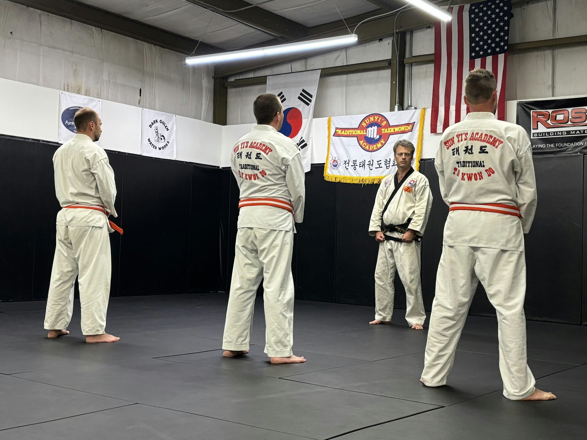 Martial arts class with four people in uniforms, banners in background, American and South Korean flags visible.