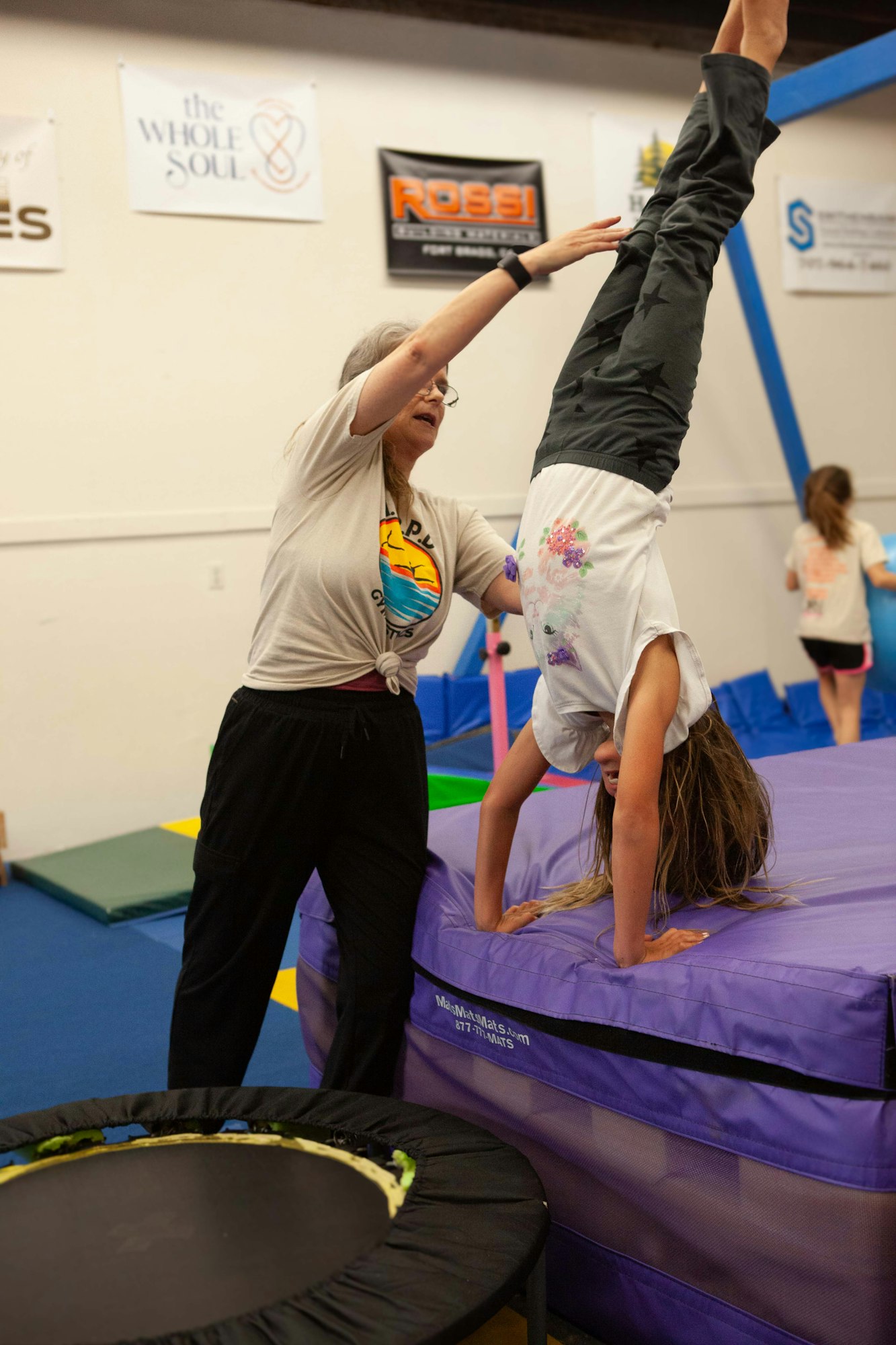 A child is doing a handstand on a gym mat while an adult assists.