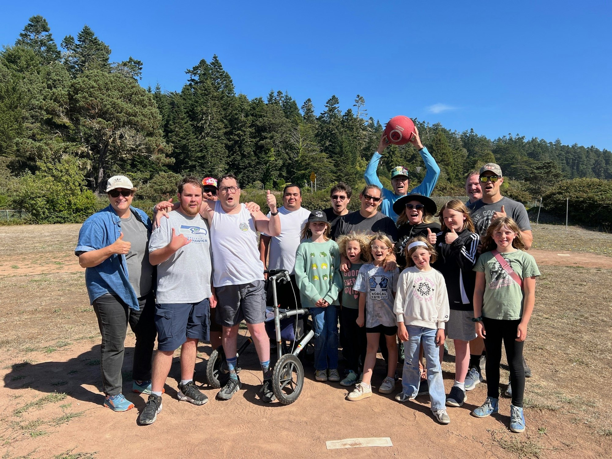 A group of people posing outdoors, some with thumbs up, one holding a football aloft. Trees and clear sky in the background.
