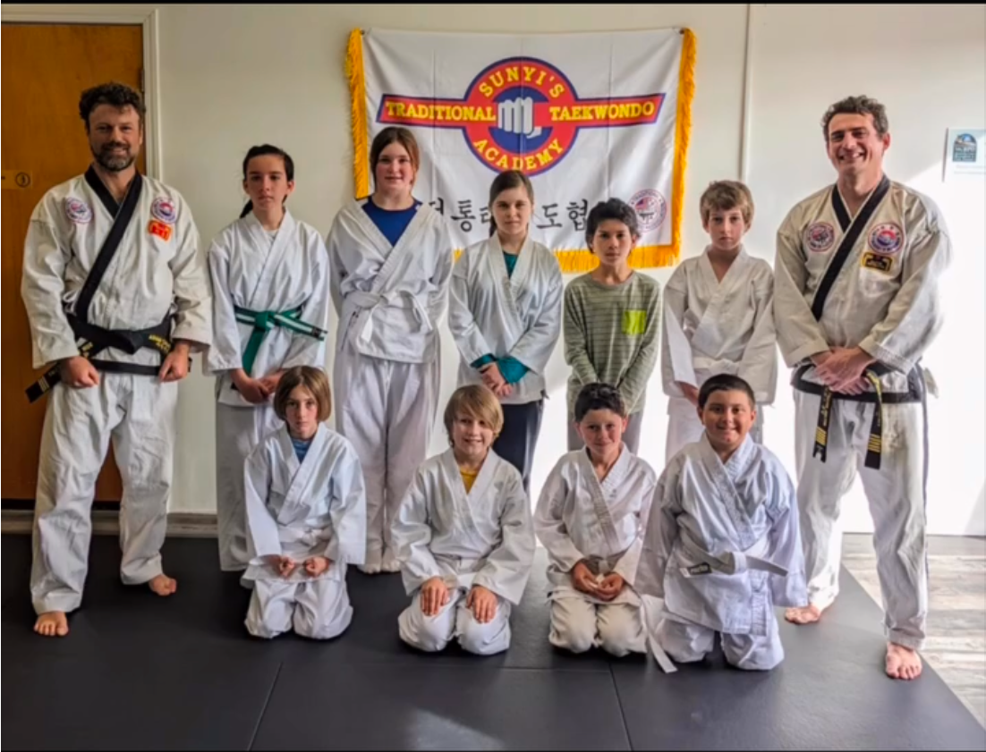 A group of people in martial arts uniforms pose in front of a "Sun Yi's Traditional Taekwondo Academy" banner.