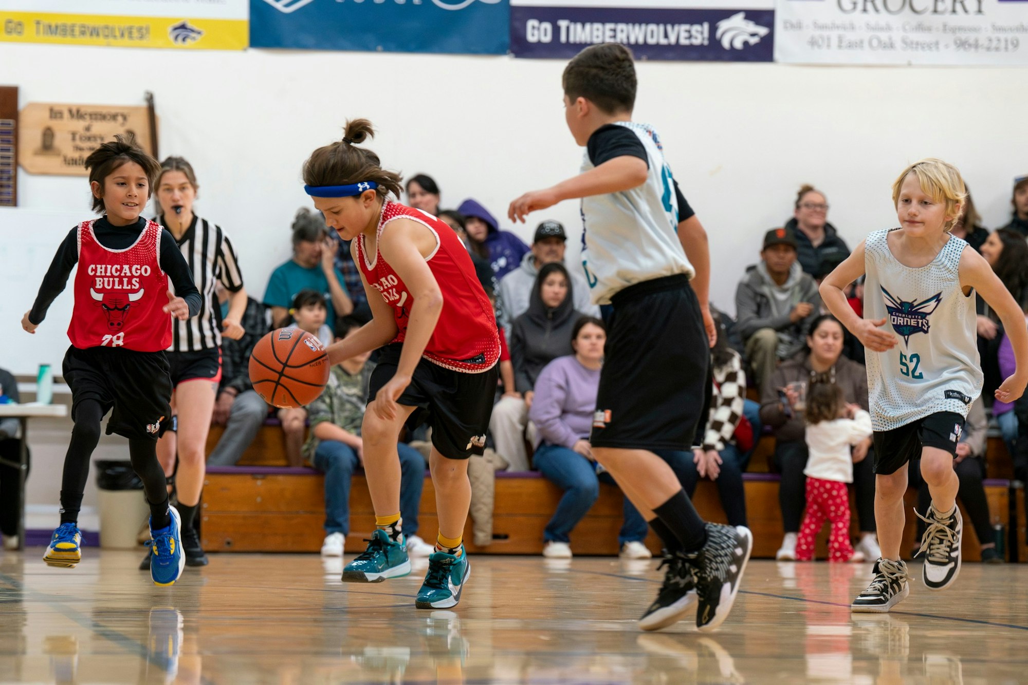 Kids playing a basketball game indoors, with spectators in the background and a referee nearby.