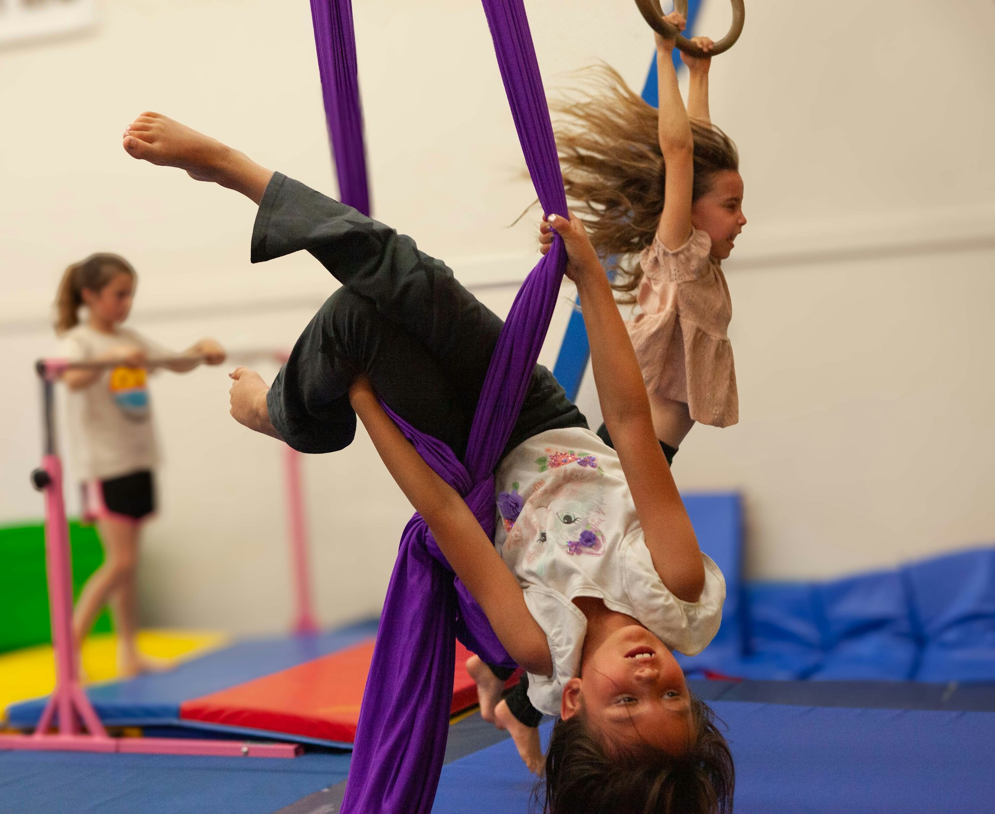 A child hangs upside down from purple aerial silks, with another child in the background, in a gymnastics studio.