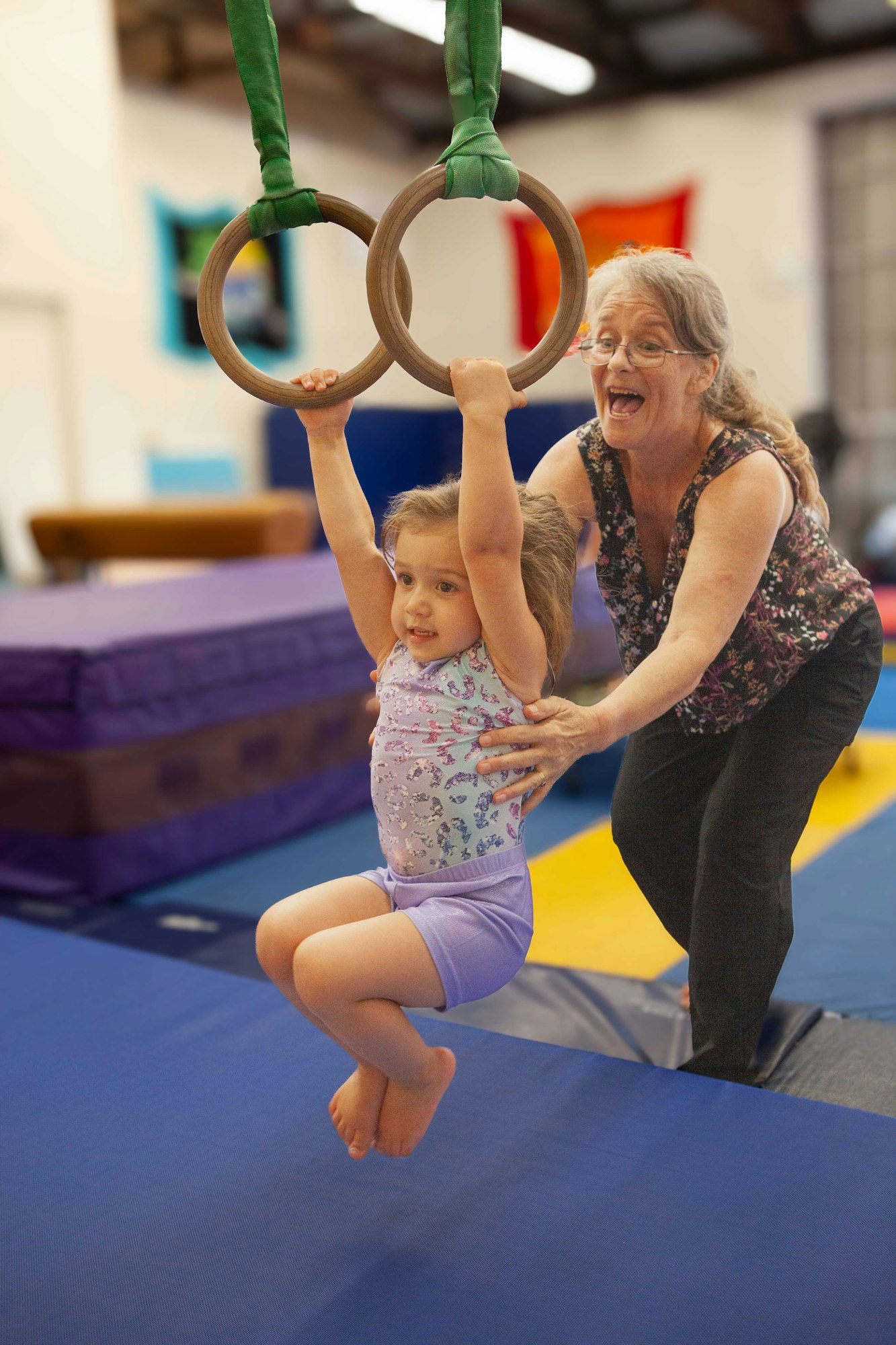 A child swinging on gymnastic rings while an adult supports her. They're in a gym with safety mats.