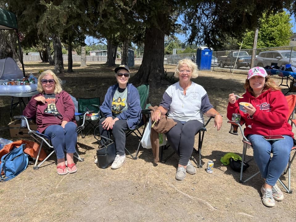 Four people sitting outdoors with a table and beverages, under shady trees, enjoying a sunny day.
