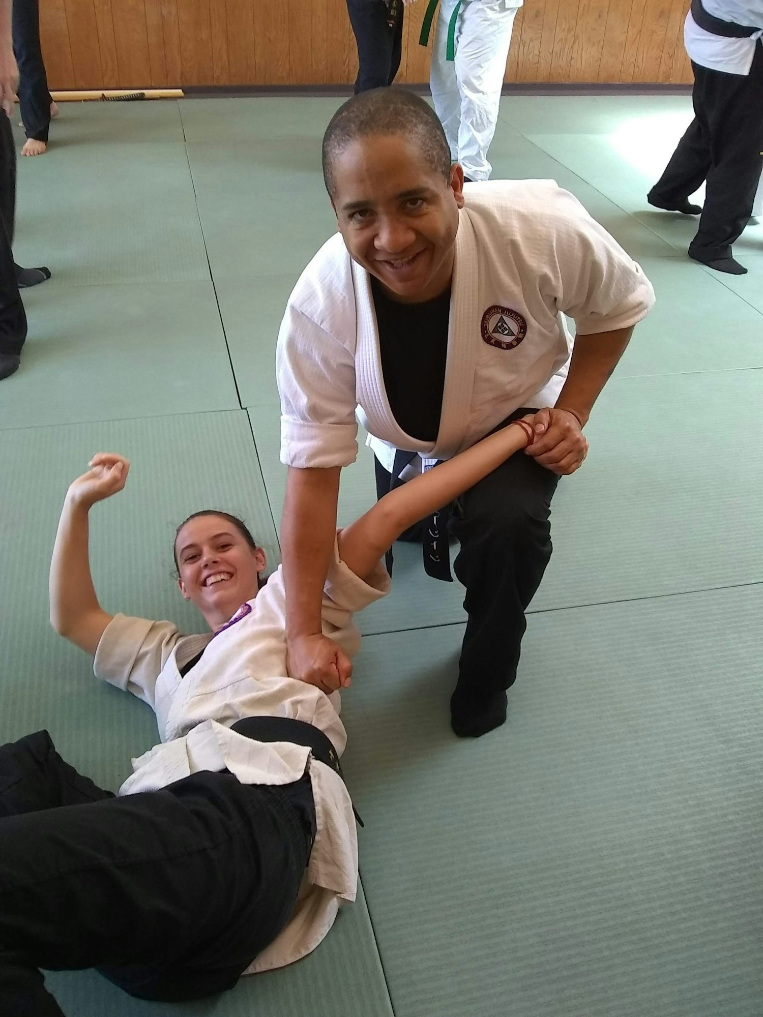 Two people in martial arts attire practicing a technique on a mat, one standing and the other on the ground smiling.