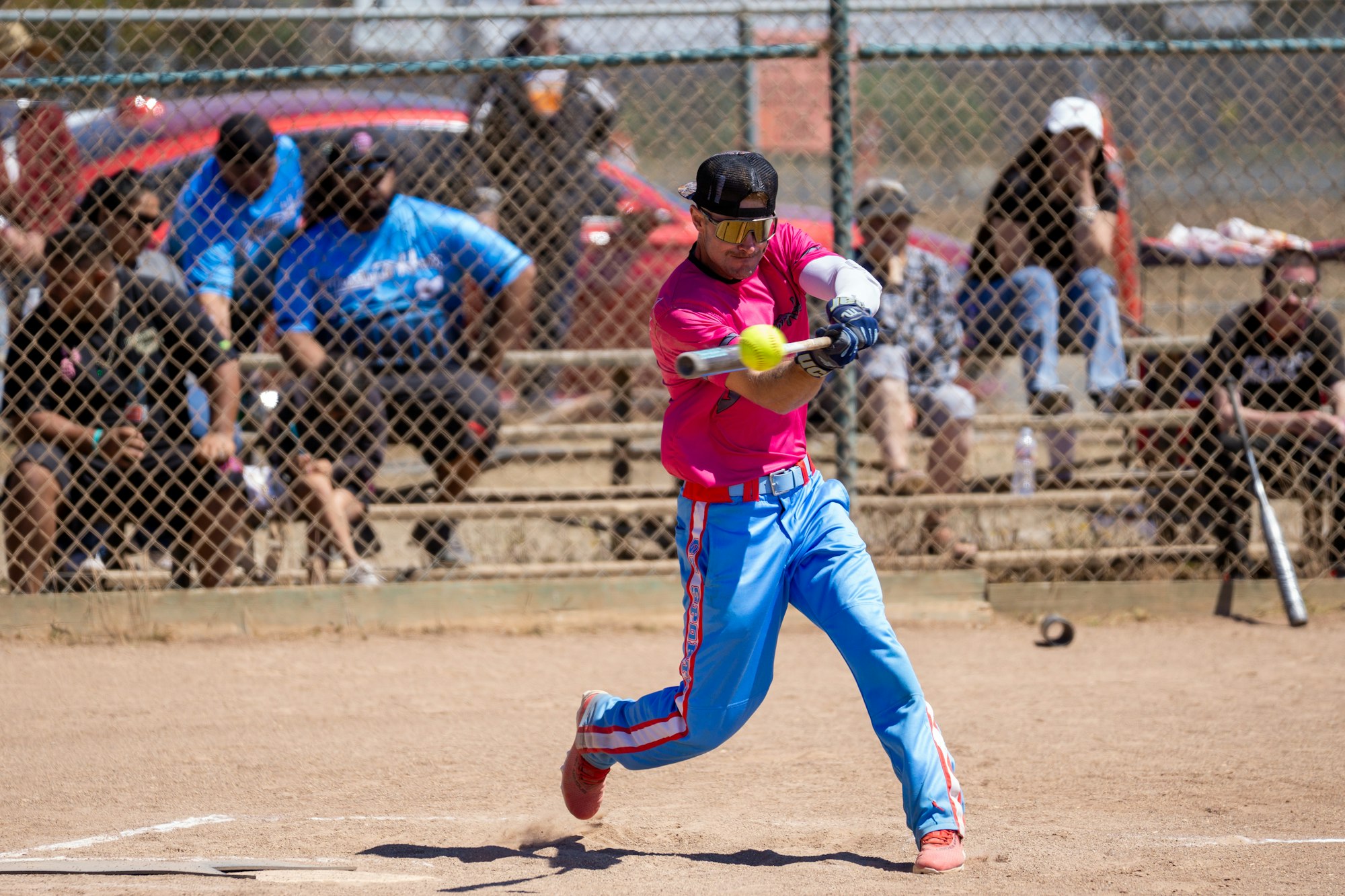 A softball player in pink and blue hits the ball, with spectators seated behind a chain-link fence.