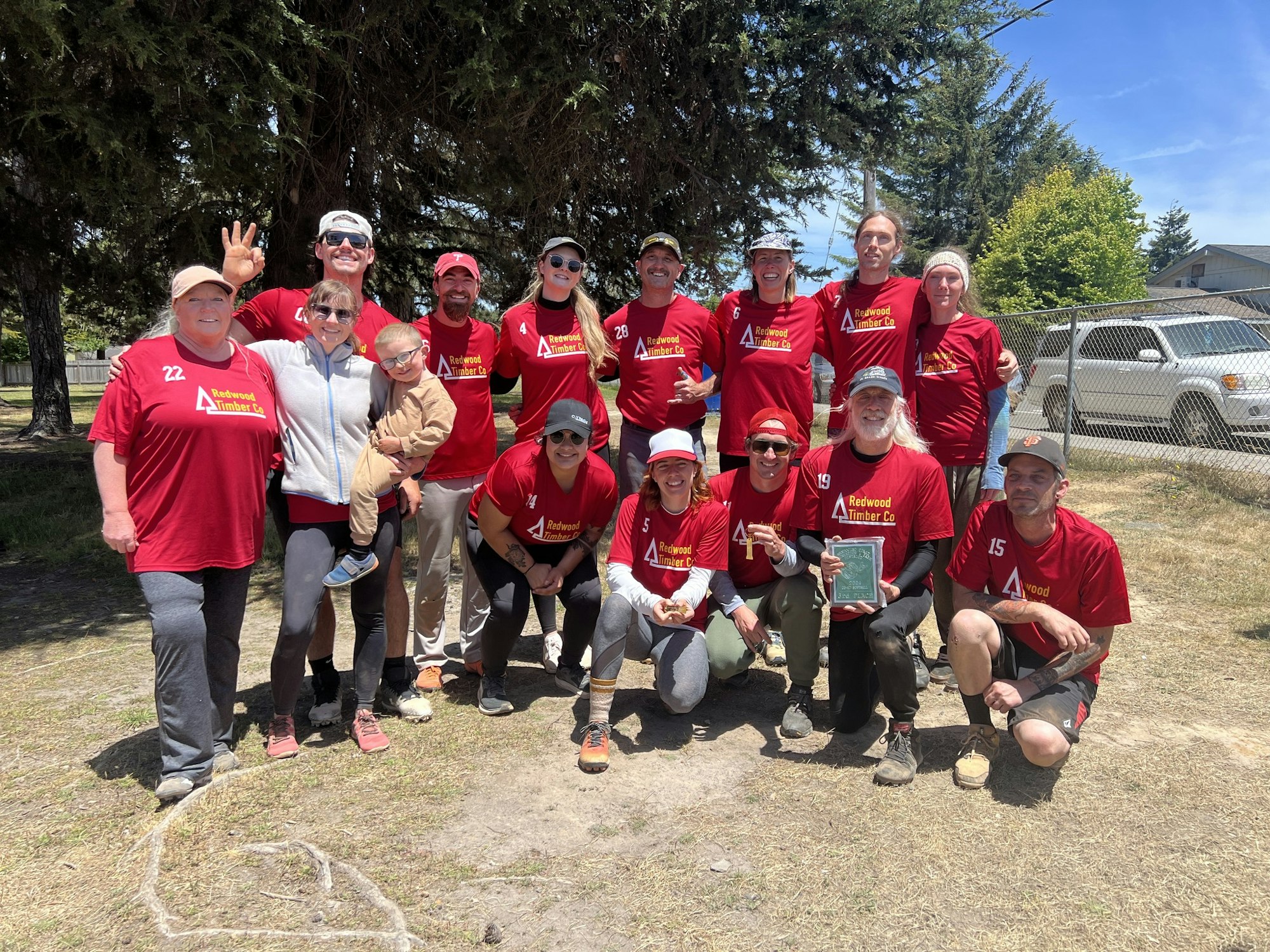 A group of smiling people in red team uniforms with a trophy, celebrating outdoors.