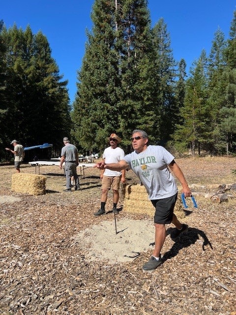 People playing horseshoes outdoors, with trees and hay bales in the background.