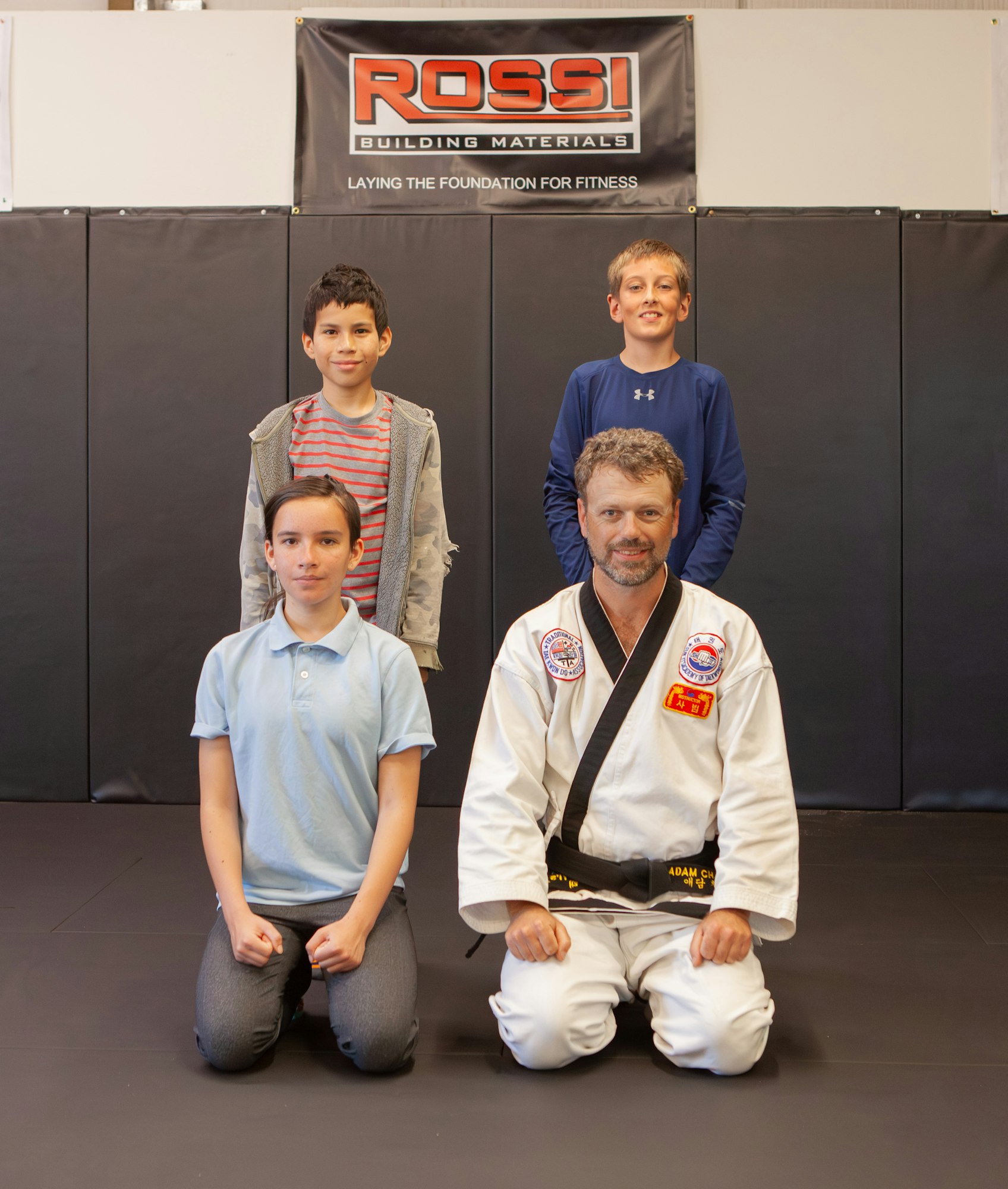 Four people posing in a martial arts dojo, one in a black belt gi, with a banner in the background.