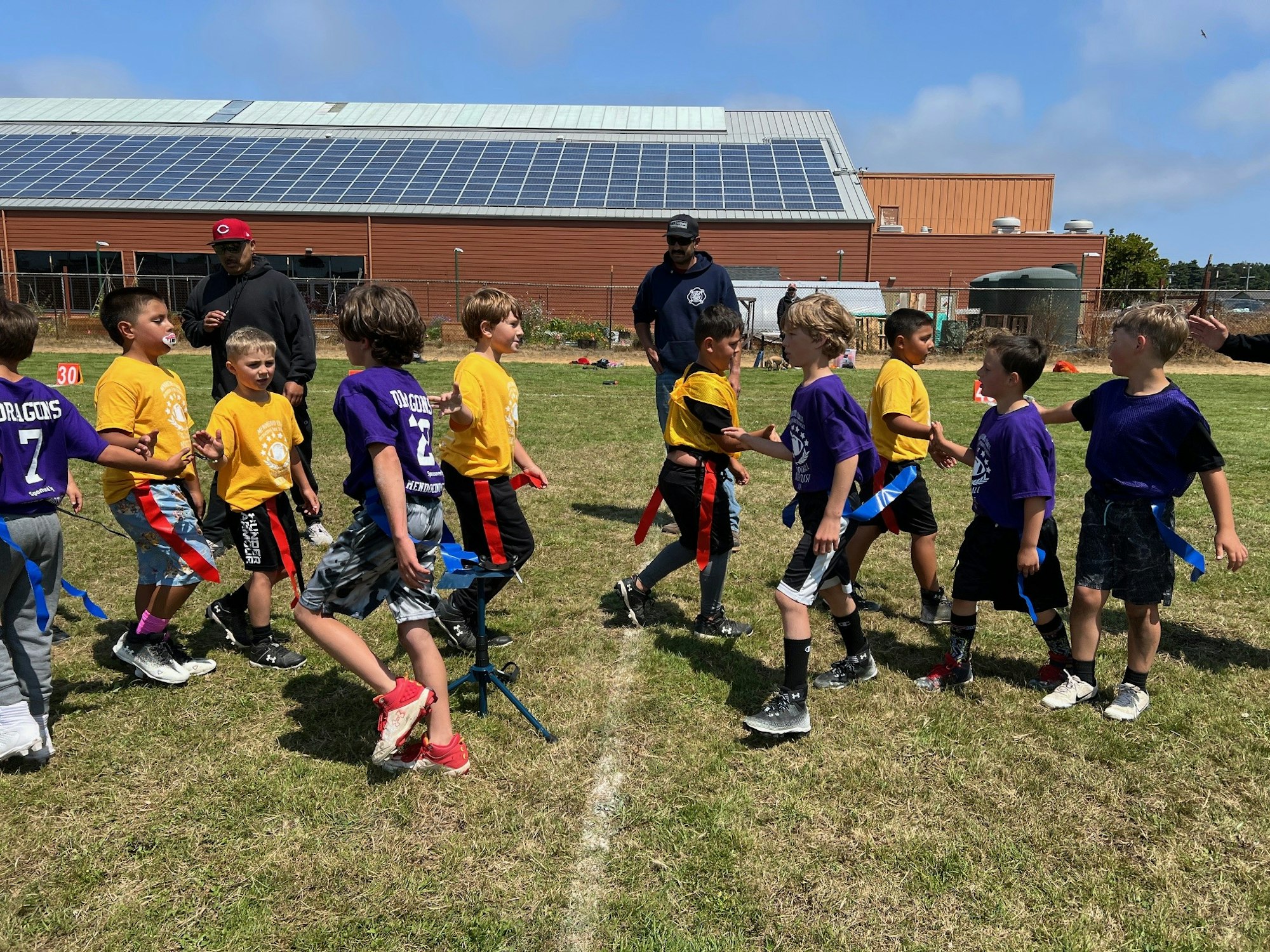Kids playing flag football on a sunny day, with two adults supervising.