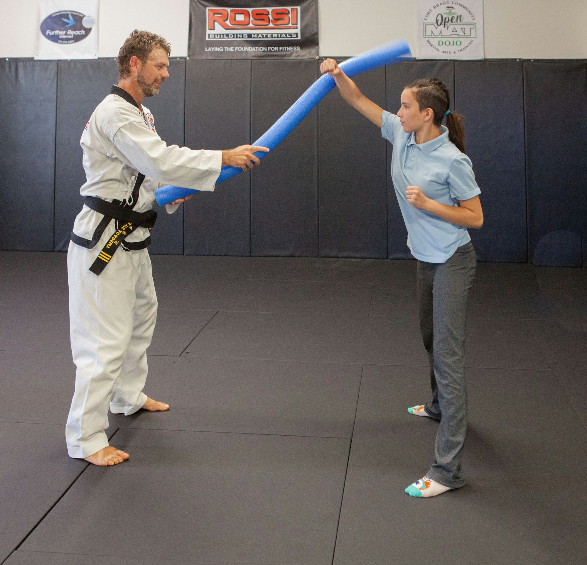 Two people are practicing martial arts using foam sticks in a training dojo.
