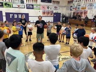 A group of children holding basketballs in a gym, listening to a coach.