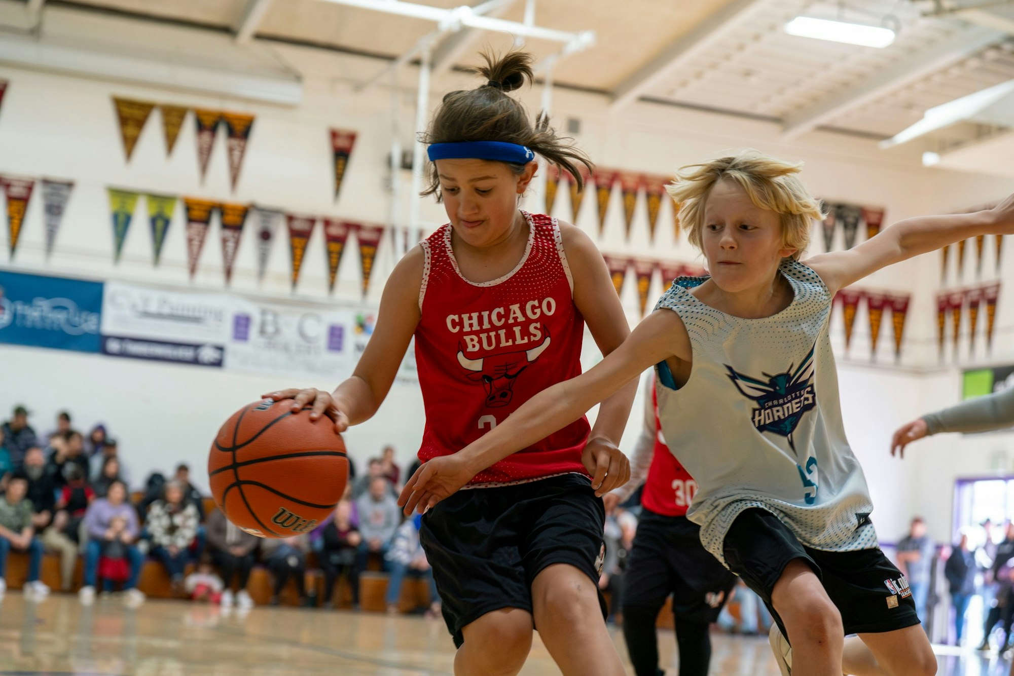 Two kids playing basketball in a gym, one in a red "Chicago Bulls" jersey dribbling past another in a "Hornets" jersey.