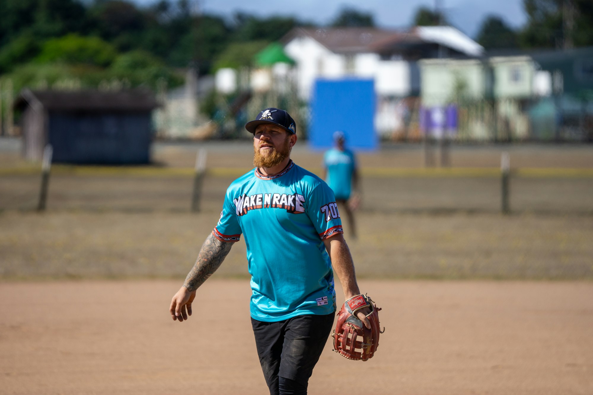 Person playing baseball, wearing a turquoise jersey, holding a glove, on a field with trees and buildings in the background.