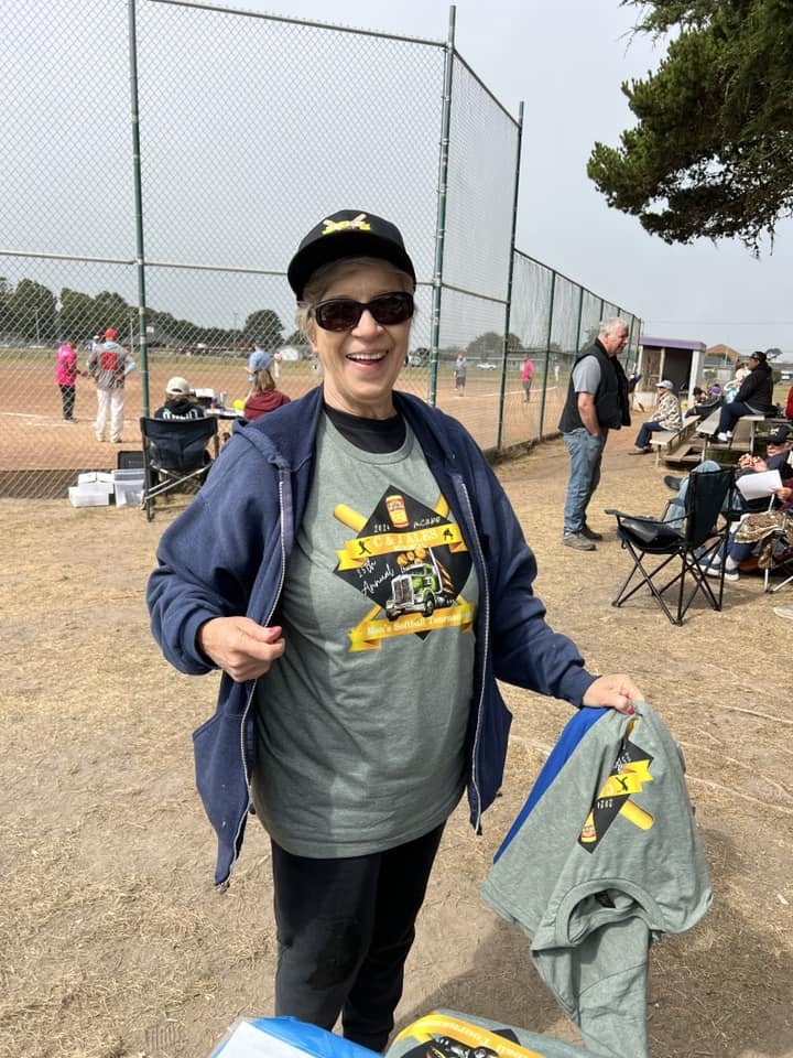 A smiling person in a baseball cap, sunglasses, and a themed T-shirt, holding clothing, at a baseball field with people in the background.