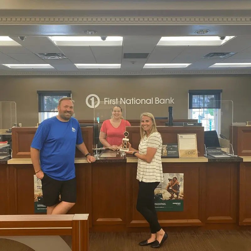 A woman holds a trophy while posing with two people at a bank, celebrating an achievement or event at First National Bank.