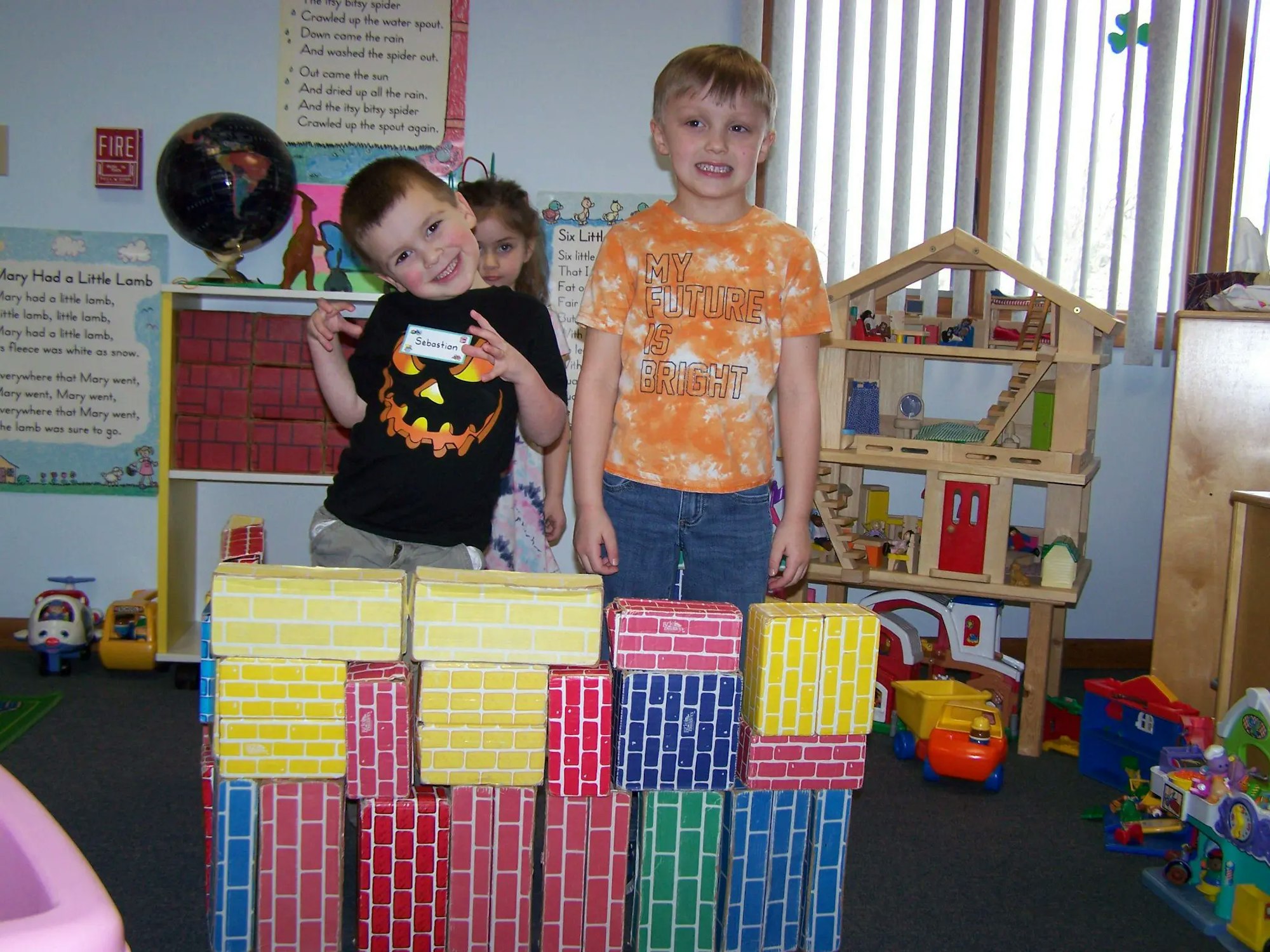 Two children pose with colorful block structures in a playroom filled with toys and educational materials.