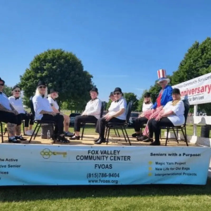 A group of people in hats and costumes sitting on chairs outdoors, celebrating at a community event for the Fox Valley Community Center.