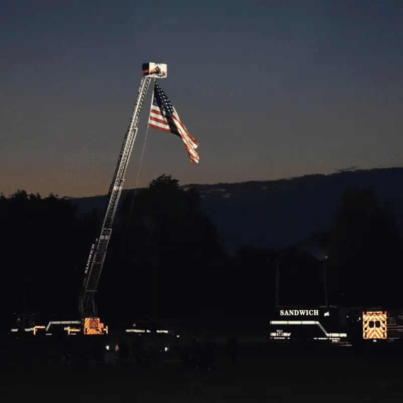 A fire truck ladder displays a large American flag at dusk, with people gathered nearby.