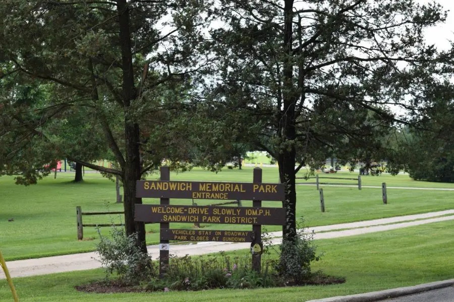 A sign at the entrance of Sandwich Memorial Park, detailing park rules and welcoming visitors. Green grassy area visible.