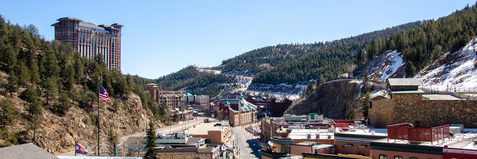 A sunny mountain town with modern buildings, American flag, light snow patches, and a forested hillside.