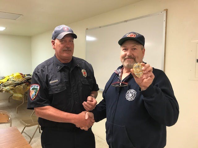 Two men are shaking hands in a room; one is a firefighter in uniform, and the other holds a badge, possibly receiving recognition.