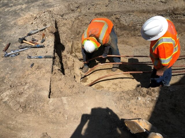 Two construction workers in safety gear work on underground pipes in a dirt trench.