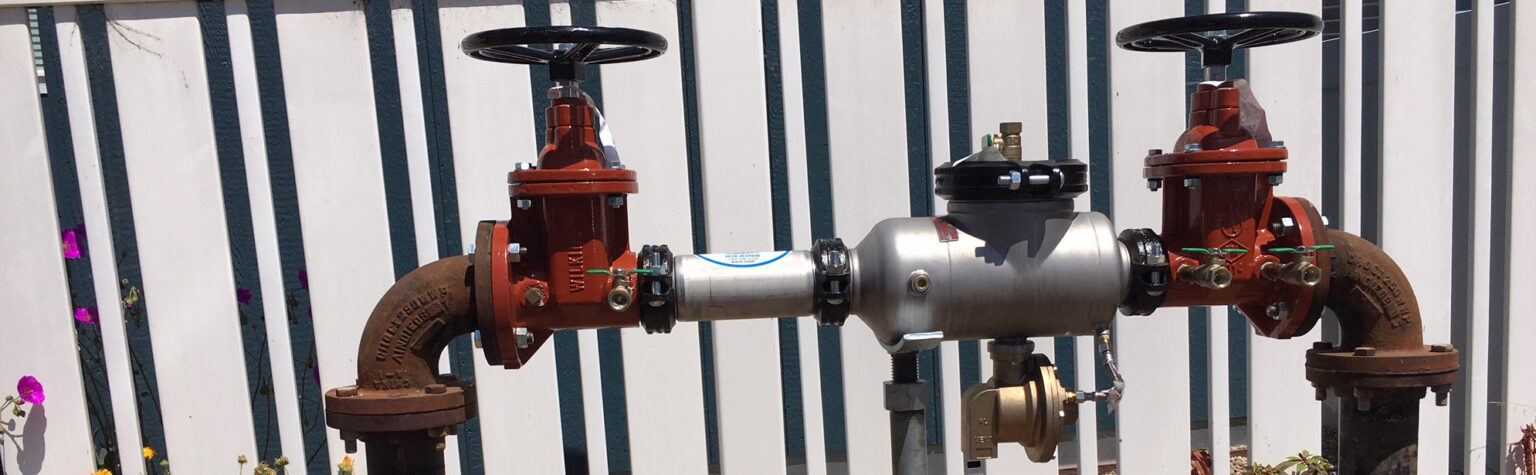 A backflow prevention device with red valve handles and metal pipes, positioned in front of a striped fence.