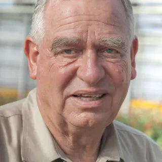 An older man with gray hair, wearing a light-colored shirt, smiles in an outdoor setting.