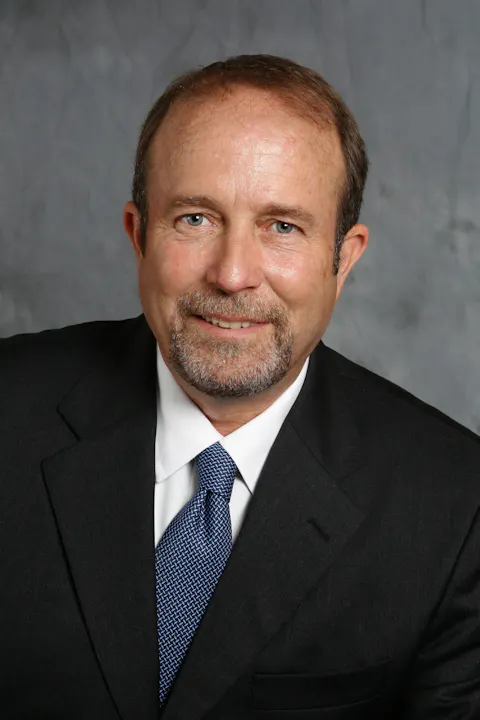 A man in a suit and tie, smiling against a gray background.