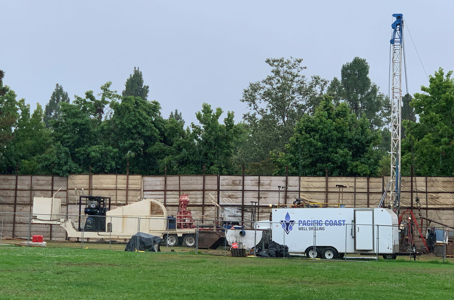 A well drilling setup with equipment and vehicles on a grassy field, surrounded by trees and a fence.