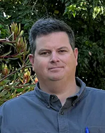 A person with short hair in a button-up shirt stands outdoors in front of greenery.