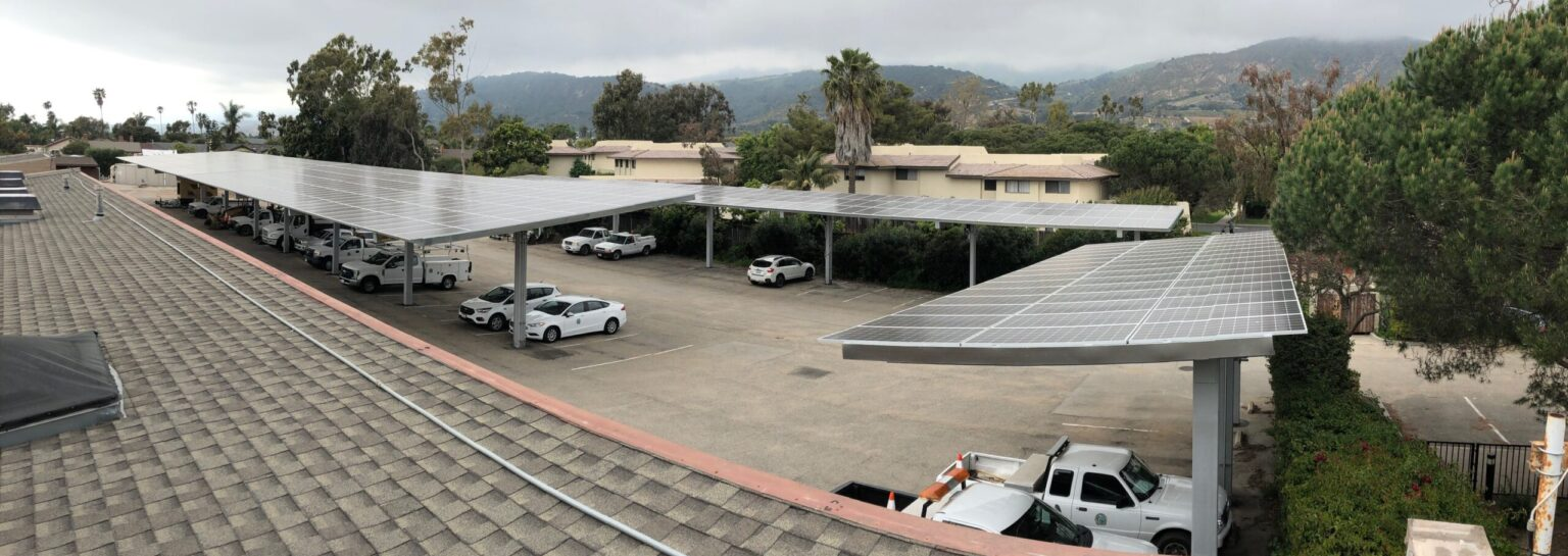 Parking lot with cars under large solar panel structures, surrounded by trees and buildings.
