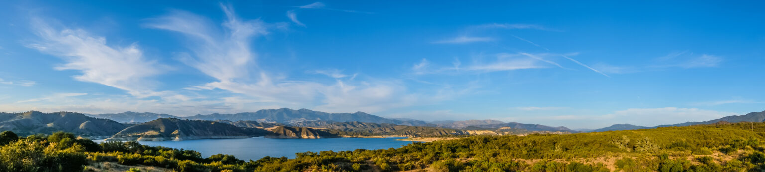 A panoramic view of a serene lake surrounded by green hills and distant mountains under a blue sky with wispy clouds.