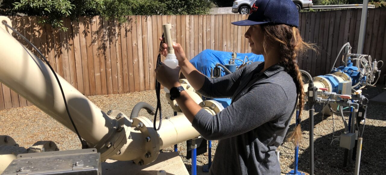 A person in a cap is inspecting equipment outdoors, holding a device beside large pipes and valves.